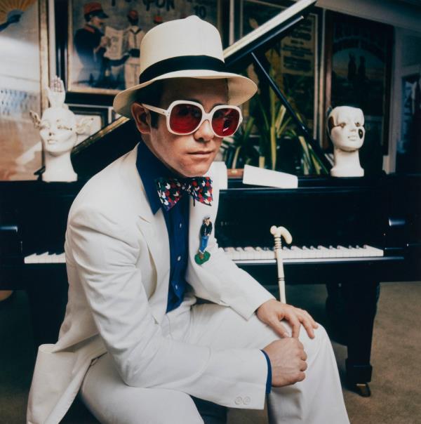 Elton John wearing a white suite, hat and large white-f<em></em>ramed sunglasses, sitting at a grand piano.